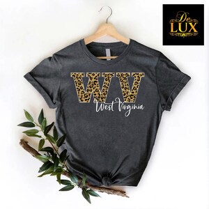 May include: Dark gray t-shirt with the letters "WW" in a leopard print design, with the words "West Virginia" in white script below. The shirt is hanging on a wooden hanger.