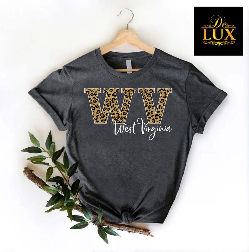 May include: Dark gray t-shirt with the letters "WW" in a leopard print design, with the words "West Virginia" in white script below. The shirt is hanging on a wooden hanger.