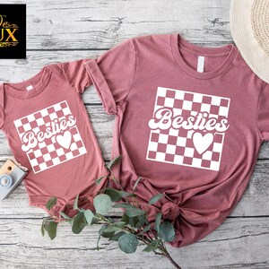 K&ouml;nnte beinhalten: Zwei mauvefarbene T-Shirts und ein Baby-Body. Jedes Kleidungsst&uuml;ck zeigt ein wei&szlig;es Schachbrettmuster mit dem Wort "Besties" und einem Herz. Der Baby-Body hat kurze &Auml;rmel, w&auml;hrend die T-Shirts kurze &Auml;rmel haben.
