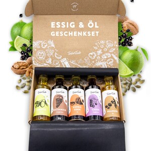 Könnte beinhalten: Ein brauner Karton mit schwarzem Deckel, geöffnet, um fünf kleine Glasflaschen mit Etiketten zu enthüllen. Die Flaschen sind in einer Reihe angeordnet und enthalten verschiedene Öle und Essige. Die Etiketten zeigen Illustrationen von Limetten, Trauben, Kürbiskernen, schwarzen Johannisbeeren und Walnüssen. Die Schachtel ist mit "ESSIG & ÖL GESCHENKSET" und "TimberTaste" beschriftet.