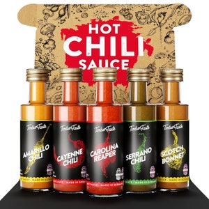 Könnte beinhalten: Ein Set aus fünf Glasflaschen mit scharfer Chilisauce, jede mit einer anderen Paprikasorte: Amarillo, Cayenne, Carolina Reaper, Serrano und Scotch Bonnet. Die Flaschen haben goldene Deckel und schwarze Etiketten mit dem "Timber Taste"-Logo. Die Verpackung enthält einen Karton mit dem Text "HOT CHILI SAUCE".