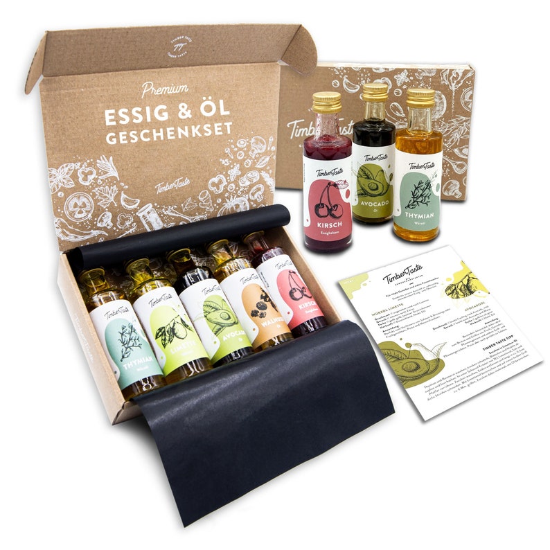 May include: A gift set of six small glass bottles of vinegar and oil in a brown cardboard box. The bottles are labeled with the words "Kirsch", "Avocado", "Thyme", "Walnut", "Blackberry", and "Cherry". The box is decorated with a white design of herbs and spices.