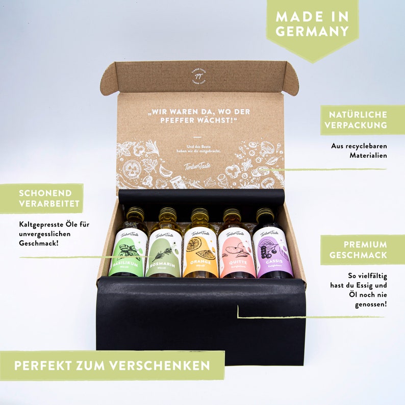 May include: A set of six small glass bottles of cold-pressed oil in a cardboard box. The bottles are labeled with different flavors, including basil, rosemary, orange, quince, and cassis. The box is decorated with a drawing of various vegetables and herbs. The text on the box reads "Wir waren da, wo der Pfeffer w&auml;chst!" and "Timber Taste".