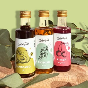 May include: Three small glass bottles with gold lids and labels. The labels are white with green, yellow, and pink accents. The bottles contain avocado oil, thyme oil, and Kirsch liqueur. The bottles are sitting on a wooden surface with green leaves.