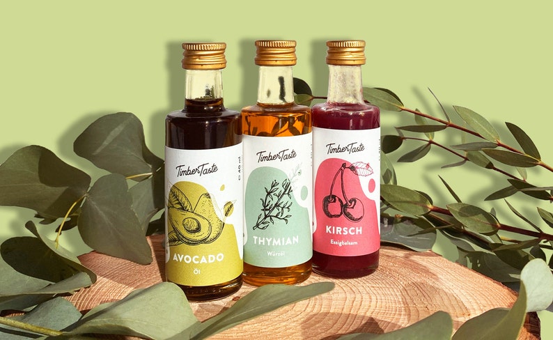 May include: Three small glass bottles with gold lids and labels. The labels are white with green, yellow, and pink accents. The bottles contain avocado oil, thyme oil, and Kirsch liqueur. The bottles are sitting on a wooden surface with green leaves.