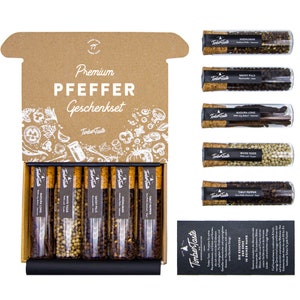 Könnte beinhalten: Ein Set aus acht Glasröhrchen gefüllt mit verschiedenen Pfeffersorten. Die Röhrchen sind mit der Pfeffersorte beschriftet, z. B. "Andaliman", "Smoky Pills" und "White Tiger". Die Röhrchen sind in einer Schachtel mit brauner Papieroberfläche und schwarzem Innenraum angeordnet. Die Schachtel ist mit "Premium Pfeffer Geschenkset" und "Timber Taste" beschriftet.
