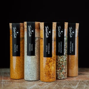 Könnte beinhalten: Sechs Glasröhren gefüllt mit verschiedenen Gewürzen, jede mit einem Korken und einem schwarzen Etikett verschlossen. Die Etiketten tragen die Aufschrift "Timber Taste" und die Gewürznamen: Paella Valencia, Tzatziki Mezedes, Chakalaka Africa, Chimichurri Argentino und Tandoori Peela Masala.