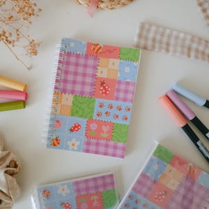 Op de afbeelding: Een spiraalgebonden notitieboek met een patchwork-ontwerp met lieveheersbeestjes, bloemen en pootafdrukken. De kaft heeft een mix van roze, blauwe en groene vierkanten. Gekleurde stiften liggen verspreid op een wit oppervlak.