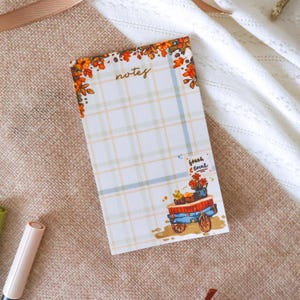 May include: A rectangular notepad featuring a light blue and yellow plaid pattern. The top border is decorated with orange and red autumn leaves. The word notes is printed at the top. A small illustration of a blue cart with flowers and the text fresh & local.