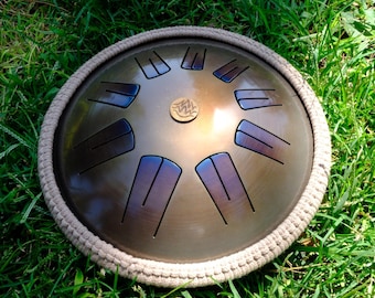 May include: A bronze-colored steel tongue drum with a circular shape and a woven rope border. The drum has several rectangular cutouts and is resting on a bed of green grass.