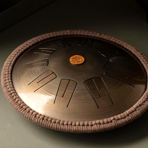 Könnte beinhalten: Eine Stahltrommel mit Bronze-Finish, geätzten Noten und einem zentralen Emblem. Die Trommel ist von einem braunen, gewebten Rand umgeben. Das Instrument ist für melodische Perkussion konzipiert.