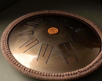 May include: A steel tongue drum with a bronze finish, featuring etched notes and a central emblem. The drum is encircled by a brown, woven border. The instrument is designed for melodic percussion.