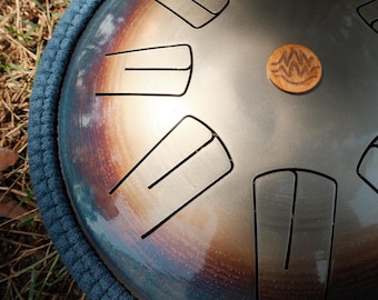 May include: A steel tongue drum with a blue padded rim. The drum has several cut-out tongues and a small wooden disc with a carved design. The drum's surface has a metallic, iridescent finish.