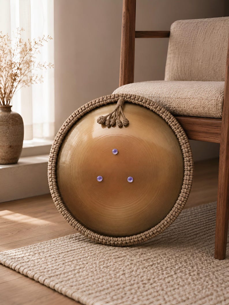 K&ouml;nnte beinhalten: Ein kreisf&ouml;rmiges Handpan-Instrument aus Messing mit einer glatten, polierten Metalloberfl&auml;che und drei kleinen, gleichm&auml;&szlig;ig verteilten kreisf&ouml;rmigen Vertiefungen. Der &auml;u&szlig;ere Rand ist mit einer dicken, geflochtenen beigen Kordel umrandet.