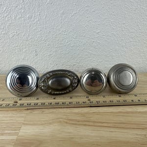 May include: Four antique metal door knobs in various designs. The knobs are silver and bronze, with one oval-shaped knob featuring an ornate design. A ruler is placed next to the knobs, showing measurements in inches.