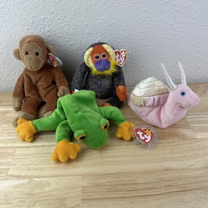 May include: Four plush toys: a brown monkey, a gray and orange orangutan, a green frog with orange feet, and a pink snail with a beige shell. Each toy has a Ty heart-shaped tag attached.