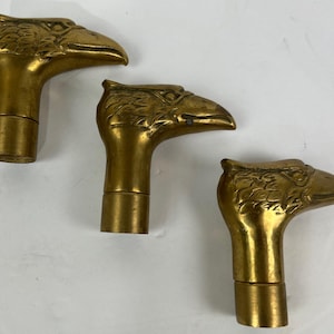May include: Three antique brass cane handles, each featuring a detailed eagle head design. The handles are a warm gold color and have a cylindrical base. The eagle heads are facing different directions.