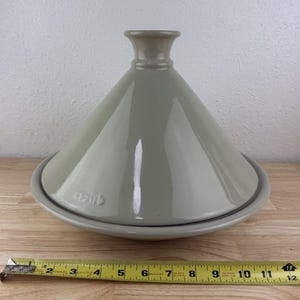 May include: A glossy light gray ceramic tagine with a conical lid and a wide circular base. The word Staub is embossed on the side of the lid. A yellow measuring tape is positioned in front, showing the base diameter is approximately 11 inches.