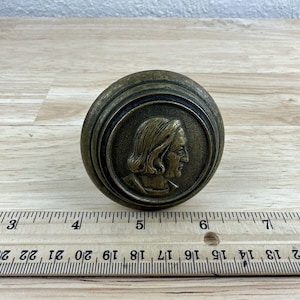 May include: A round, brass door knob with a raised, detailed profile of a person's face in the center.