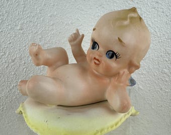 May include: A vintage porcelain figurine of a cherubic baby, lying on a yellow pillow. The baby has rosy cheeks, blue eyes, and a small tuft of blonde hair. The figurine is a collectible item.