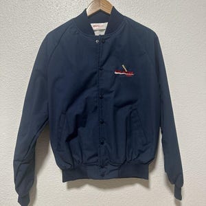 May include: A navy blue bomber jacket with a snap closure and a red, white, and blue embroidered logo that says "The Great American Sportsman".