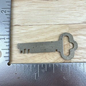May include: A vintage metal key with a decorative bow-shaped handle and a series of teeth. The key is placed on a wooden surface next to a metal ruler. The key's length is approximately 2 inches.
