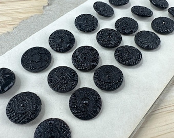 May include: A collection of black, round buttons with intricate, raised patterns. The buttons are arranged on a piece of aged, torn paper, suggesting a vintage or antique aesthetic. The buttons vary slightly in size and design, adding to their unique appeal.