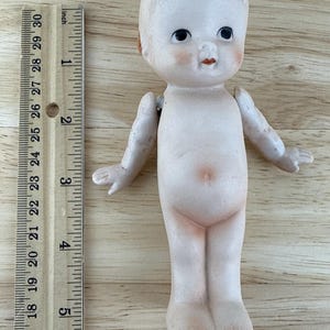 May include: A vintage bisque doll with a painted face, rosy cheeks, and red lips. The doll has articulated arms and legs. A wooden ruler with inch measurements is next to the doll.