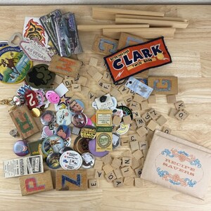 May include: A collection of vintage ephemera, including patches, pins, wooden letter tiles, and a recipe holder. The patches feature various designs, including a Las Vegas sign and a conservation logo. The wooden tiles are for a game.