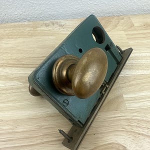 May include: An antique door lock with a green metal plate and a brass-colored oval doorknob. The lock includes a metal strike plate and is secured with screws. The item is placed on a wooden surface.