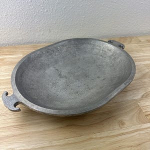 May include: Oval-shaped, gray metal serving dish with textured surface and two decorative handles. The dish is made of a heavy metal and sits on a wooden surface. The dish is likely used for serving food.