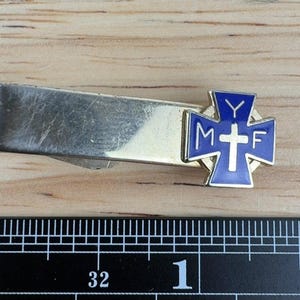 May include: A gold-tone metal tie clip featuring a blue enamel cross emblem with the letters M, Y, and F and a central white cross. A black ruler with white markings is positioned below, showing the clip is approximately 1.5 inches long.