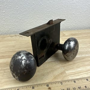 May include: An antique door lock set with two round, metal doorknobs and a rectangular lock mechanism. The metal has a distressed, aged appearance. A wooden ruler is in the foreground, providing scale. The set is likely for a vintage or rustic door.