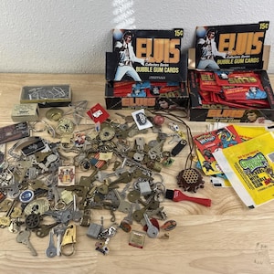 May include: A collection of assorted metal keys on rings, two boxes of Elvis Collectors Series bubble gum cards, and a stack of colorful stickers. The Elvis boxes feature a black and yellow design with a 15 cent price tag and red card packs.