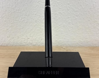 May include: A black pen on a black stand with the word "SHEAFFER" printed on the front. The stand has a ruler along the bottom edge. The pen is a sleek, modern design.