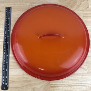 May include: Round, orange enameled cast iron pot lid with a red rim and handle. A black ruler is placed next to the lid for scale. The lid is approximately 12 inches in diameter.