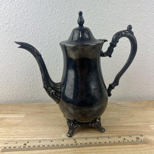 May include: A silver-colored coffee pot with a curved spout and ornate handle. The pot has a lid with a decorative finial and rests on three small feet. A ruler is placed in front of the pot.