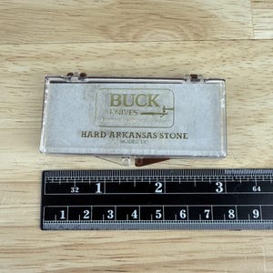 May include: A clear plastic case containing a Buck Knives Hard Arkansas Stone, Model 130. The case has a hinged lid and a brown label with the Buck Knives logo and text. A black ruler with white markings is below the case.