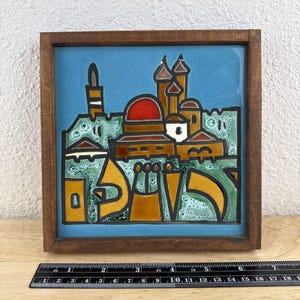 May include: A framed ceramic tile featuring a colorful illustration of a Middle Eastern cityscape. The artwork includes buildings with red and brown domes, a tall tower, and text in a yellow and orange hue against a blue background. The frame is made of wood.