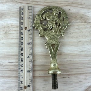 May include: A vintage metal decorative piece with a cherub design, measuring approximately 6 inches in length. The piece has a threaded base and a detailed cherub figure within an ornate frame. The metal has a tarnished, antique finish.