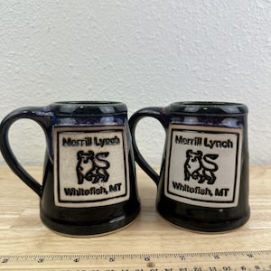 May include: Two black ceramic mugs with handles. Each mug features a white rectangular emblem with the text "Merrill Lynch" above a stylized animal and the text "Whitefish, MT". The mugs are on a wooden surface.
