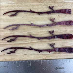 May include: Four decorative, twig-shaped items with a dark burgundy color and a textured finish. Each has a branch-like design with small offshoots and a rounded end. They are arranged on a light wood surface.