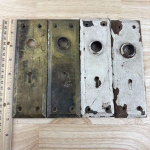 May include: Four antique door plates. Two are brass, one with a keyhole and the other with a round opening. The other two are painted white with chipped paint, one with a keyhole. A ruler is placed next to the plates.
