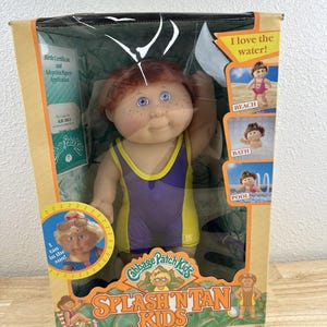 May include: Cabbage Patch Kids Splash 'N Tan doll in original packaging. The doll has red hair, blue eyes, and is wearing a purple and yellow swimsuit. The box features images of the doll at the beach, bath, and pool.