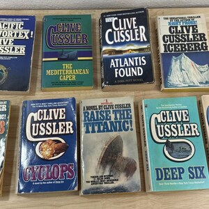 May include: A collection of nine paperback books by Clive Cussler, including titles such as Pacific Vortex!, The Mediterranean Caper, Atlantis Found, Iceberg, Vixen 03, Cyclops, Raise the Titanic!, Deep Six, and Treasure, all featuring illustrated covers.