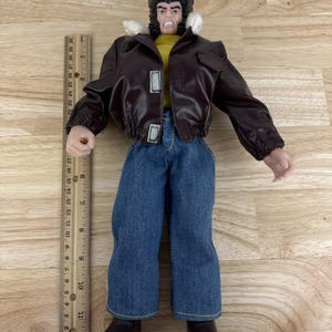 May include: A Wolverine action figure wearing a brown leather jacket, blue jeans, and black boots. The figure has a yellow shirt and a stern expression. A ruler is placed next to the figure, showing measurements in inches.