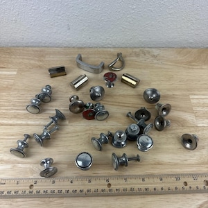 May include: A collection of vintage metal cabinet knobs and pulls in various shapes and sizes. The hardware includes silver-toned knobs, some with decorative details, and a few brass-toned pieces. A wooden ruler is placed near the hardware.