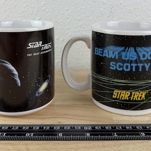 May include: Two white ceramic Star Trek mugs. One mug features a spaceship graphic and the text "Star Trek The Next Generation". The other mug has the phrase "Beam Us Down Scotty" and the Star Trek logo.