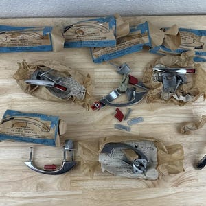 May include: An assortment of vintage car door handles and related parts, some wrapped in brown paper. The handles are chrome with red and white accents. Several blue and tan boxes are also present, likely containing additional components.