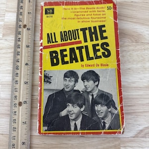 May include: A vintage book titled "All About The Beatles" by Edward De Blasio. The cover features a photo of the Beatles and text on a yellow and red background. The book is next to a wooden ruler.
